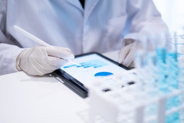 A lab technician in gloves reviews data charts on a tablet beside racks of sample tubes.